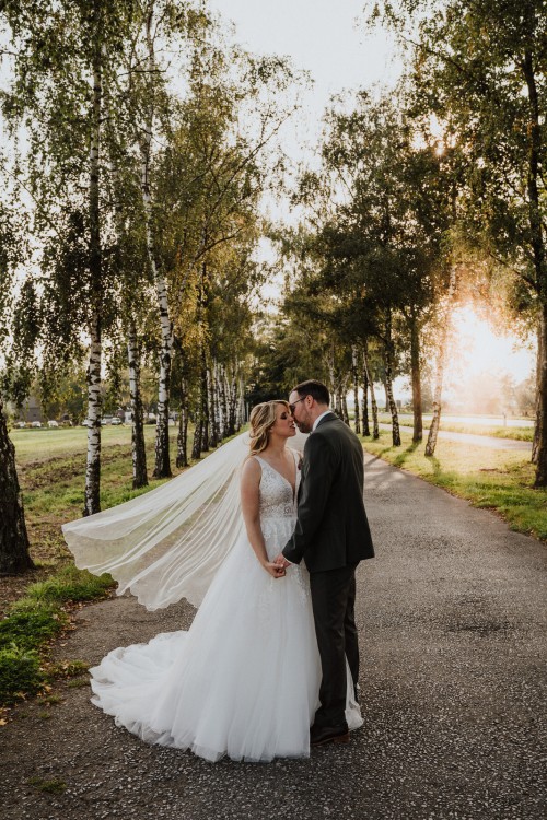 Ein Brautpaar küsst sich mitten in einer Birkenallee beim Sonnenuntergang.